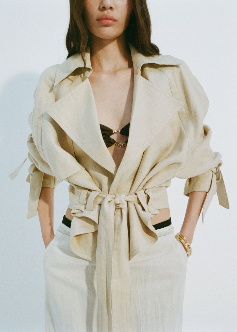 A woman poses in a studio wearing a belted trench coat, light pants, and gold bracelets. The shot is a medium shot with a white background.