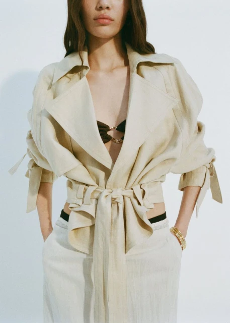 A woman poses in a studio wearing a belted trench coat, light pants, and gold bracelets. The shot is a medium shot with a white background.