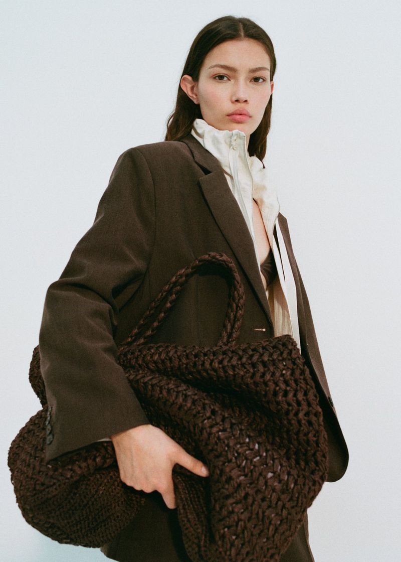 Brunette woman wearing a brown suit and holding a dark brown knit bag against a plain white background in a studio shot.