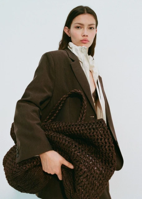 Brunette woman wearing a brown suit and holding a dark brown knit bag against a plain white background in a studio shot.