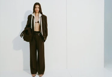 Model wearing a brown blazer suit and white hoodie carrying a dark woven bag against a plain white studio wall