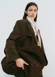 Model wearing a brown blazer holding a large dark woven handbag against a plain white background for Mango.