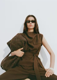 Model wearing dark sunglasses and a brown sleeveless belted top, holding a woven dark brown bag against a plain white background.
