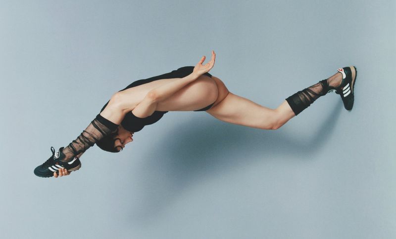 A person in a black bodysuit and sheer socks is floating horizontally in front of a light blue background, holding a black Adidas sneaker.