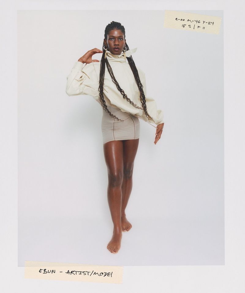 A dark-skinned model with braids poses in front of a white background wearing a neutral-toned top and skirt, a fashion studio shot.