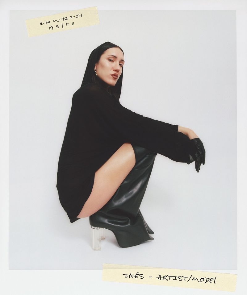 Portrait of a dark-haired woman posing in black clothing and black boots with clear heels against a white backdrop in a photo studio.