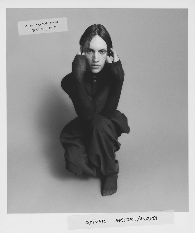 Black-and-white portrait of a person in a dark outfit, kneeling with hands near their face against a neutral background in a photo studio.