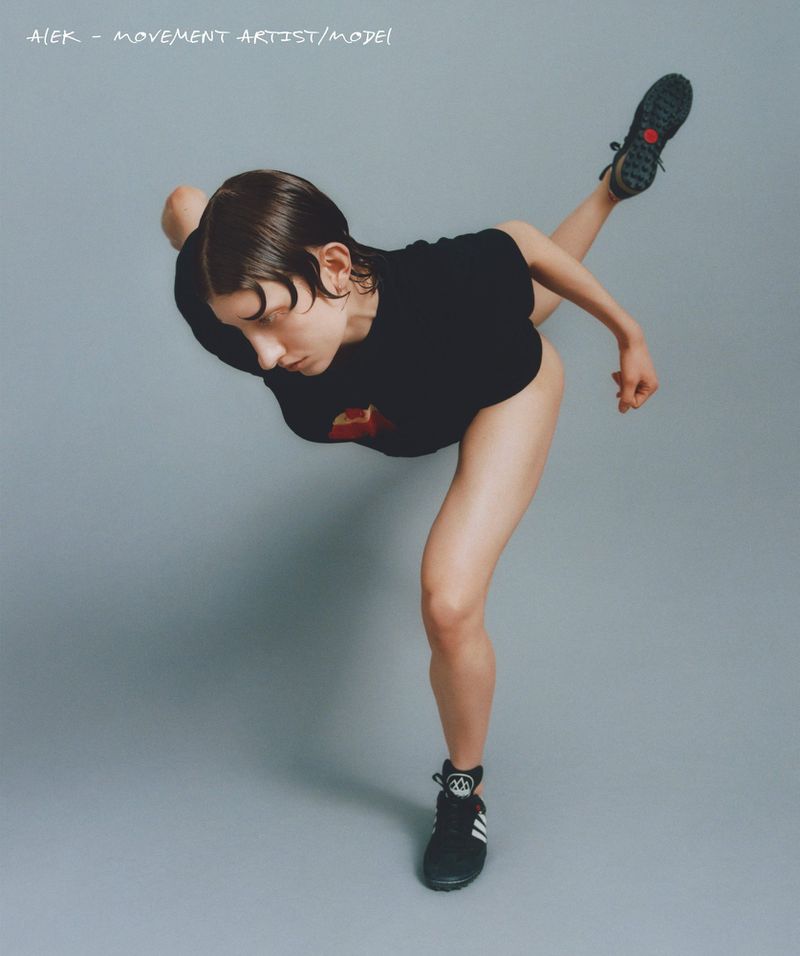 Woman in black shirt and athletic shoes performs a dance move against a grey backdrop, one leg extended backward.