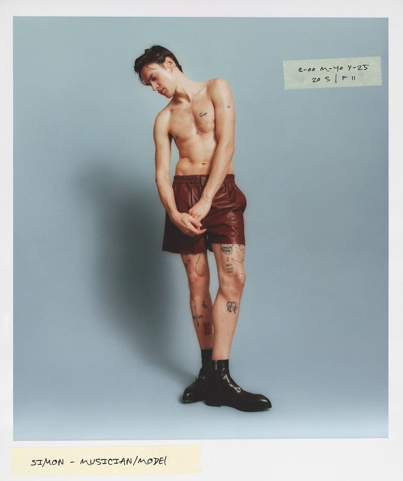 A shirtless slender adult male in brown shorts and black boots poses in a full shot against a light blue background, with handwritten notes.