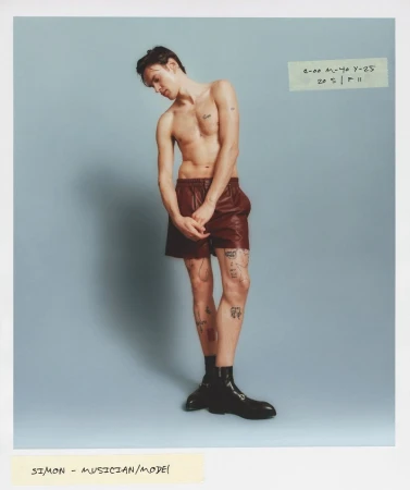 A shirtless slender adult male in brown shorts and black boots poses in a full shot against a light blue background, with handwritten notes.