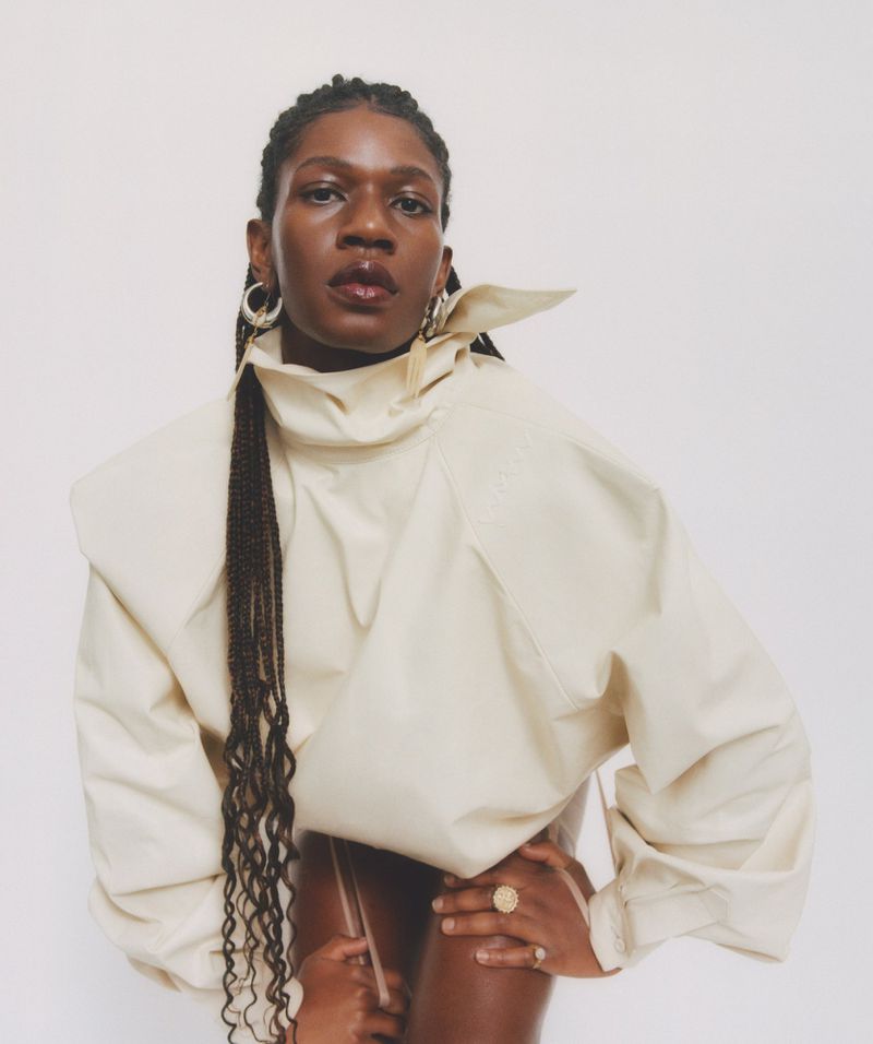 Portrait of a stylish African-American woman wearing a sculptural cream-colored top with braided hair and hoop earrings against a white background.