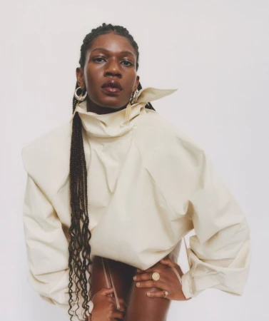 Portrait of a stylish African-American woman wearing a sculptural cream-colored top with braided hair and hoop earrings against a white background.