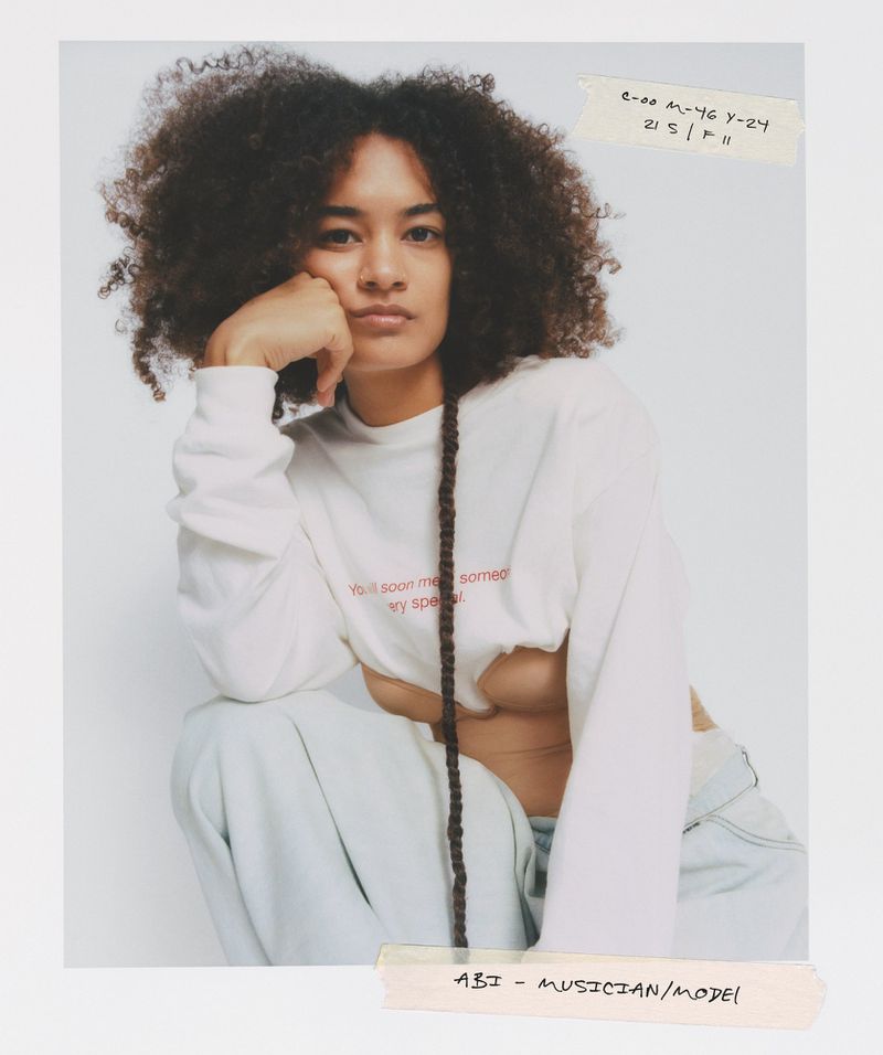 Portrait of a young adult woman with curly afro hair and long braid, wearing a white long-sleeve shirt, posing in a photo studio.