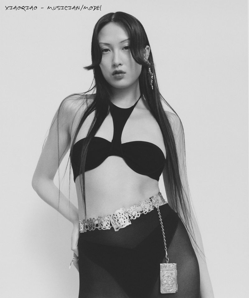 Monochrome portrait of an Asian female model in a fashion bra top, ornamented belt, and long black hair against a white backdrop.