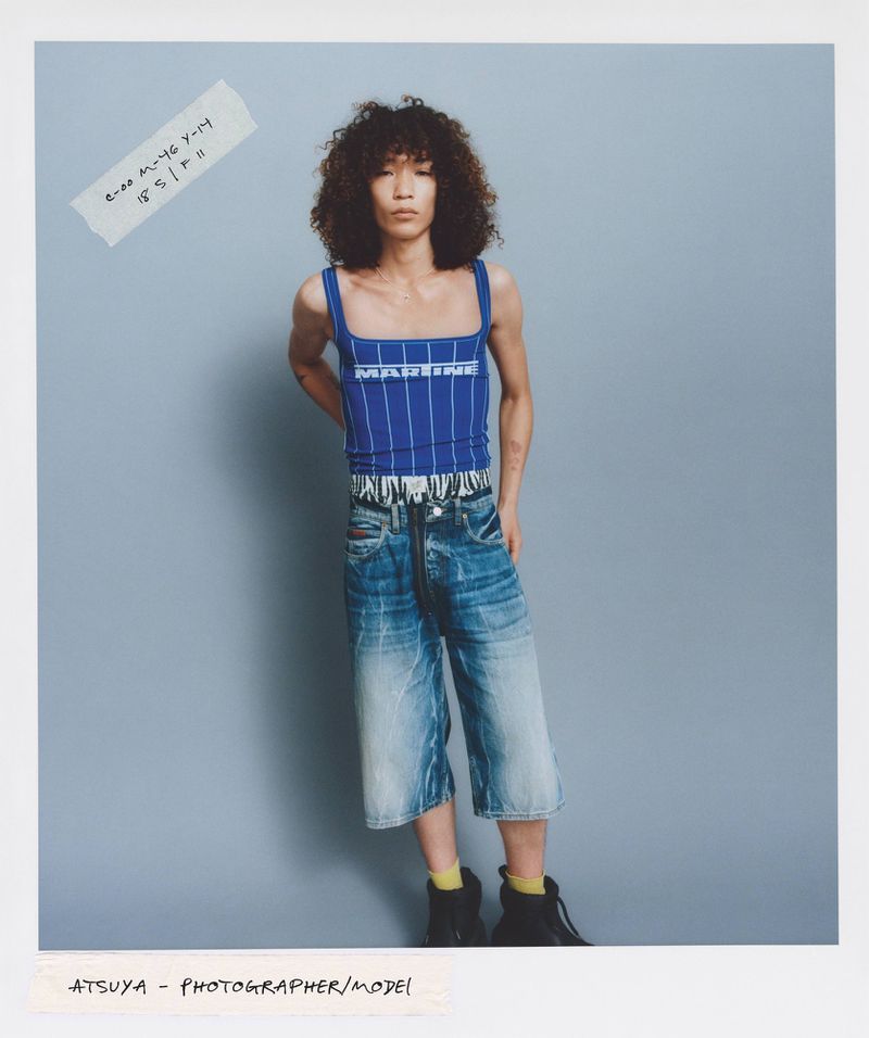 Fashion shot of a young adult with curly hair wearing a blue and white top with blue jean shorts and black boots against a blue background.