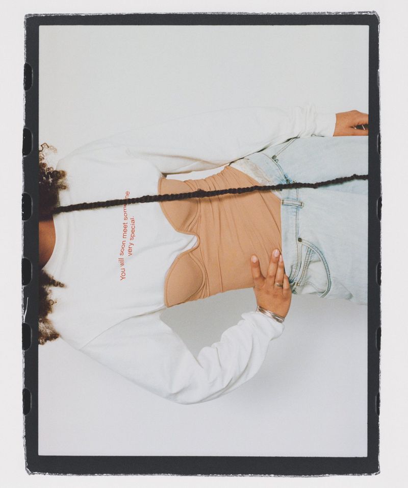 A woman with curly hair poses while wearing a beige top, light-blue denim, and a white, long-sleeved sweatshirt against a white background.