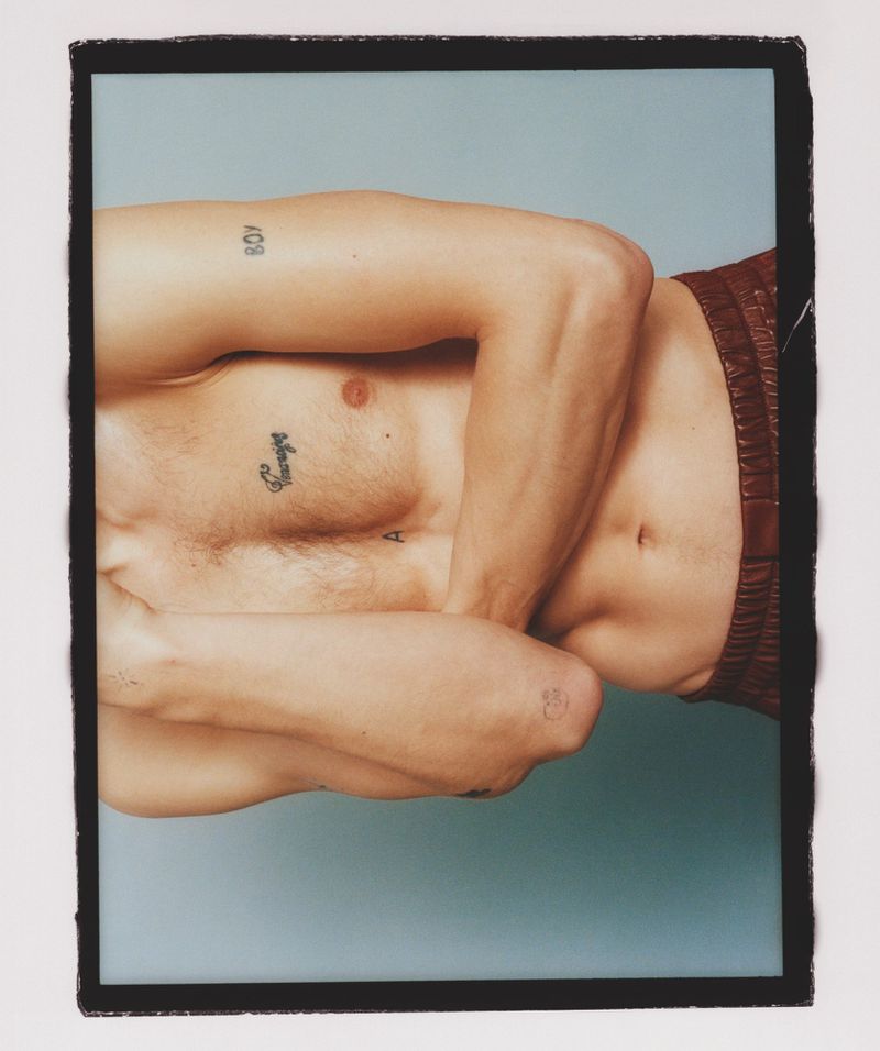Shirtless adult male poses with brown shorts against a blue backdrop, showcasing arm tattoos and a relaxed studio setting.