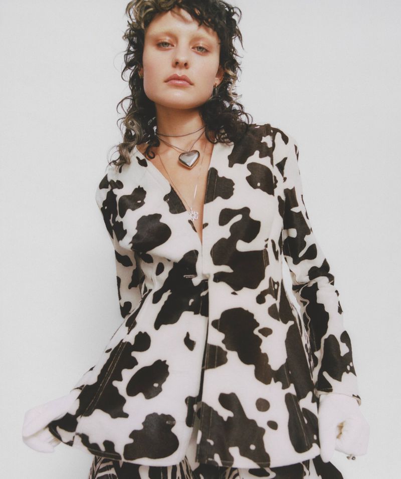 Portrait of a person with a mullet hairstyle wearing a cow-print jacket with a heart necklace against a white studio background.