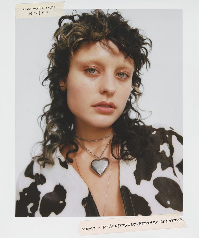 Close-up studio portrait of a young woman with curly dark hair and a cow-print shirt, accessorized with a heart pendant necklace.