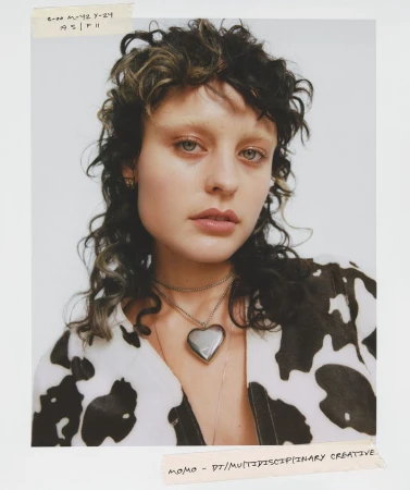 Close-up studio portrait of a young woman with curly dark hair and a cow-print shirt, accessorized with a heart pendant necklace.