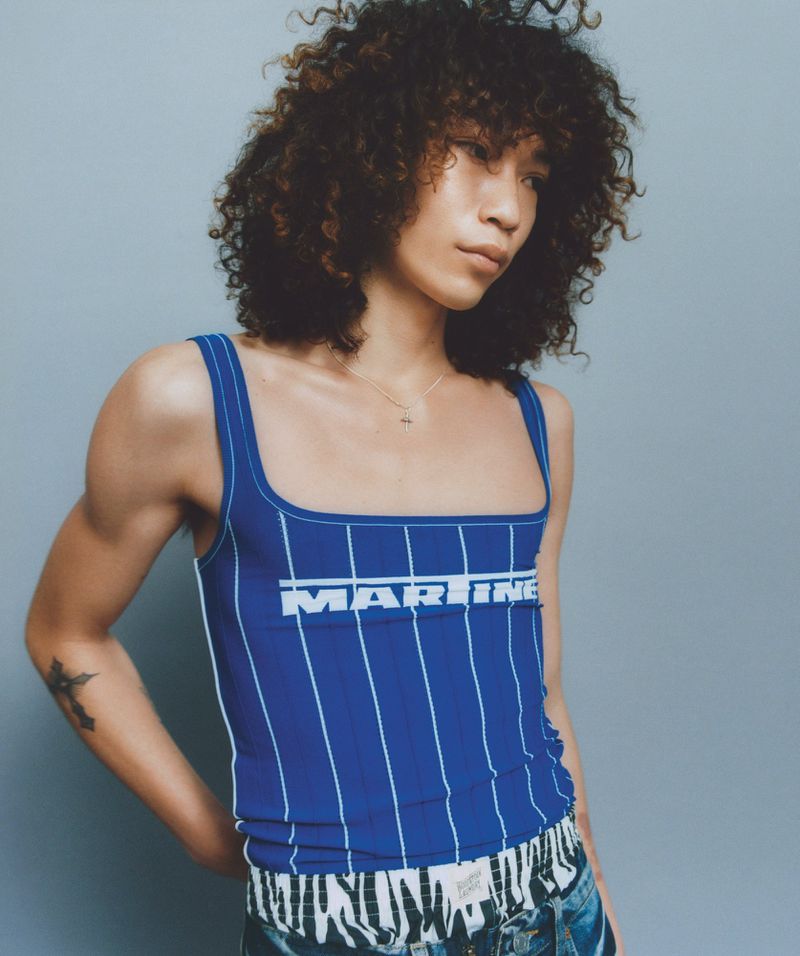 A person poses in a blue and white striped tank top with curly hair, set against a neutral backdrop in a fashion photography context.