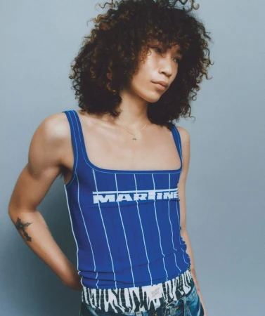 A person poses in a blue and white striped tank top with curly hair, set against a neutral backdrop in a fashion photography context.