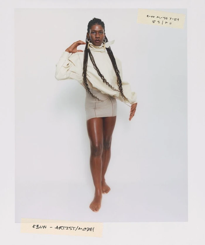 Black woman with long braids wearing a cream top and fitted skirt against a white background