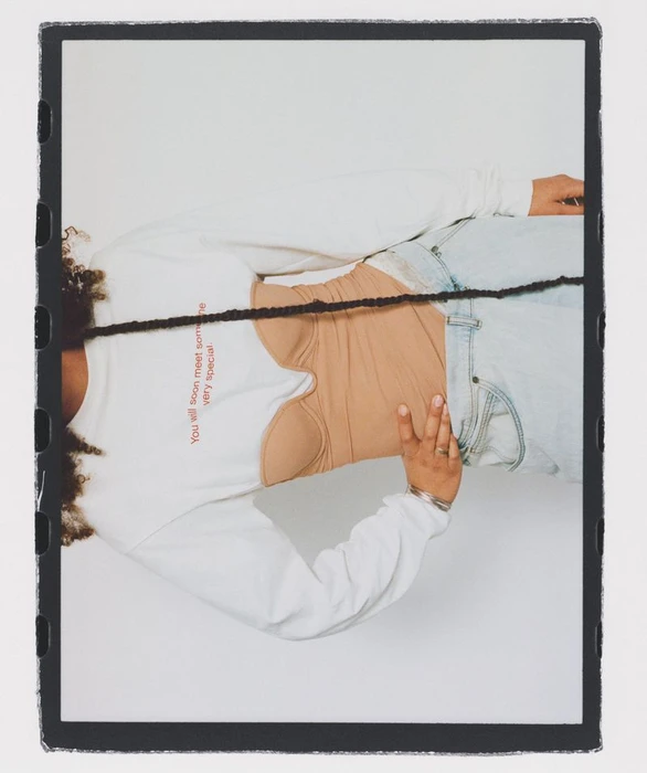 Close-up portrait of a person wearing a white long-sleeve top, nude bustier, and light denim jeans against a white background.