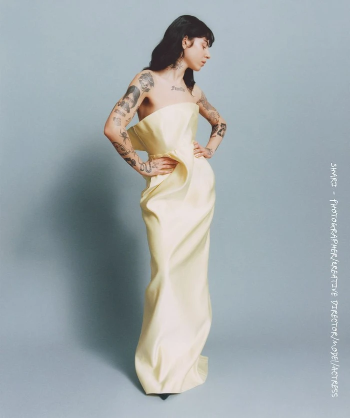 Tattooed woman wearing a strapless pale yellow gown posing against a grey studio backdrop for Hube Magazine editorial.