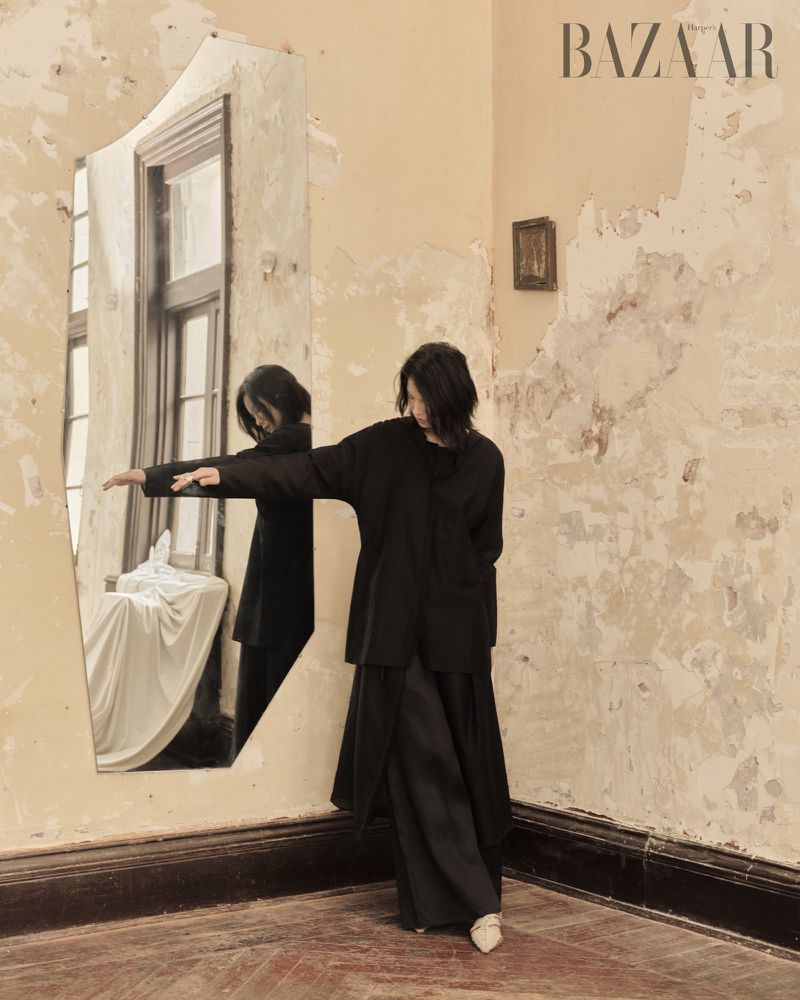Woman in black clothing stands in corner near broken mirror in decayed interior, captured in a fashion editorial style with textured walls.