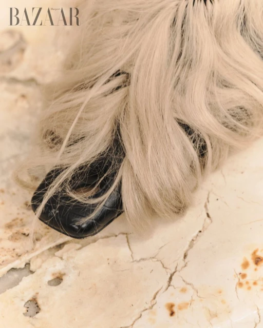 Close-up of a black shoe adorned with blonde hair extensions, set against a cracked, textured surface, and featuring the Bazaar logo.