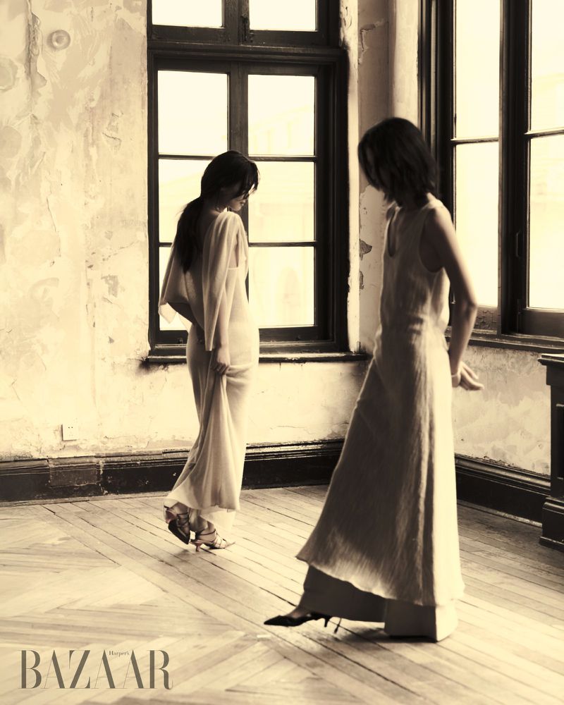 Two women pose in long dresses in an old room with large windows and worn walls, vintage style. Fashion editorial and indoor scene.