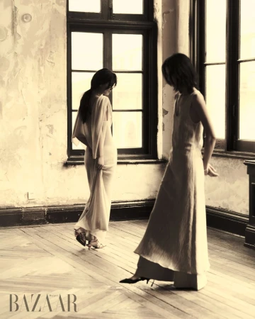 Two women pose in long dresses in an old room with large windows and worn walls, vintage style. Fashion editorial and indoor scene.