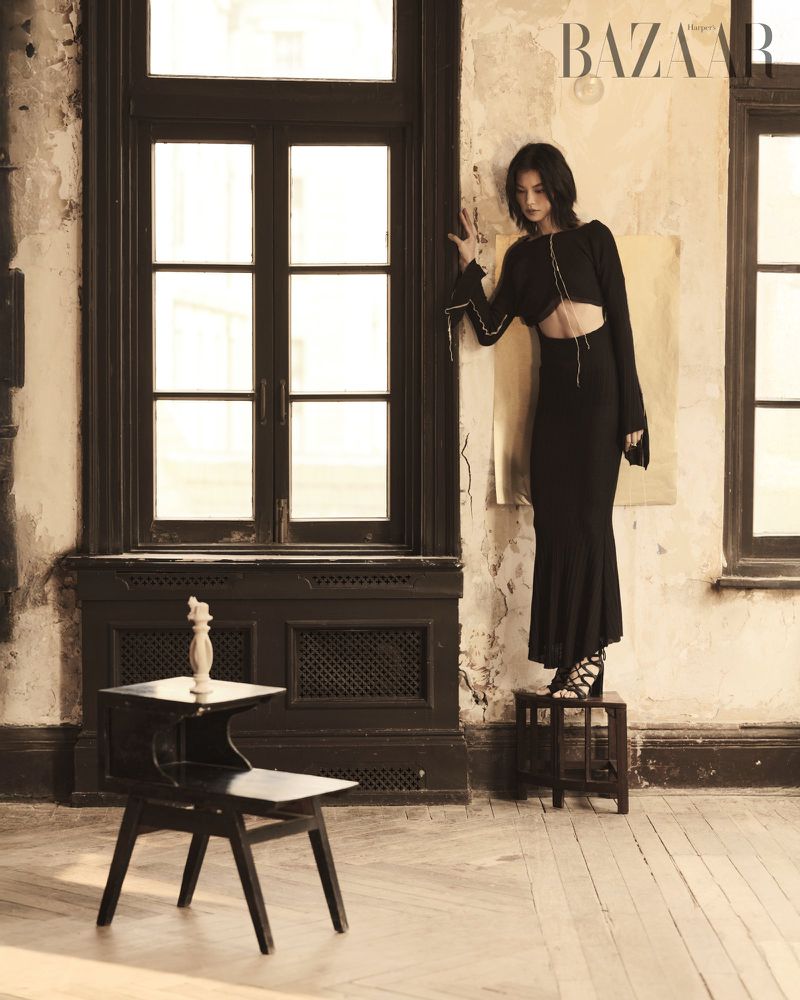 A young woman poses in a black outfit inside a room with old windows and walls. The scene features a vintage fashion style with sepia tones.