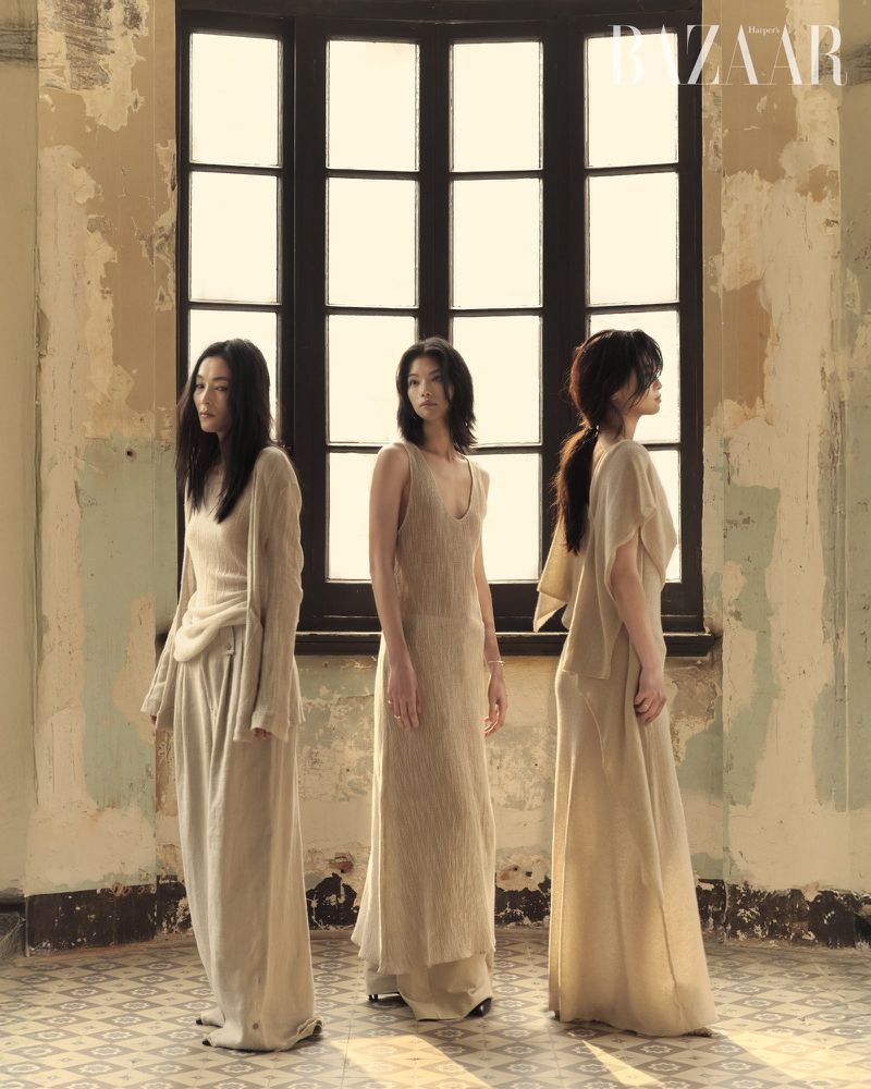 Three Asian women pose in long beige dresses in front of a window in a fashion editorial, set in an old building with vintage details.