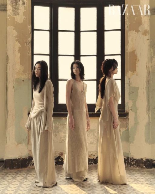 Three Asian women pose in long beige dresses in front of a window in a fashion editorial, set in an old building with vintage details.
