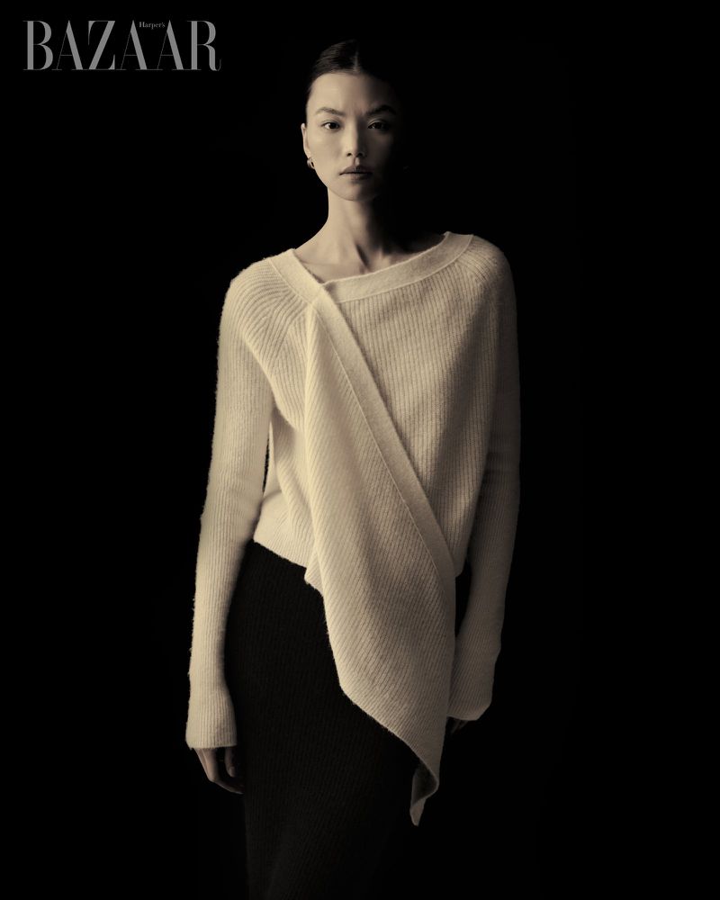 Elegant studio portrait of an Asian woman in a cream-colored sweater and black skirt against a dark background, fashion editorial.