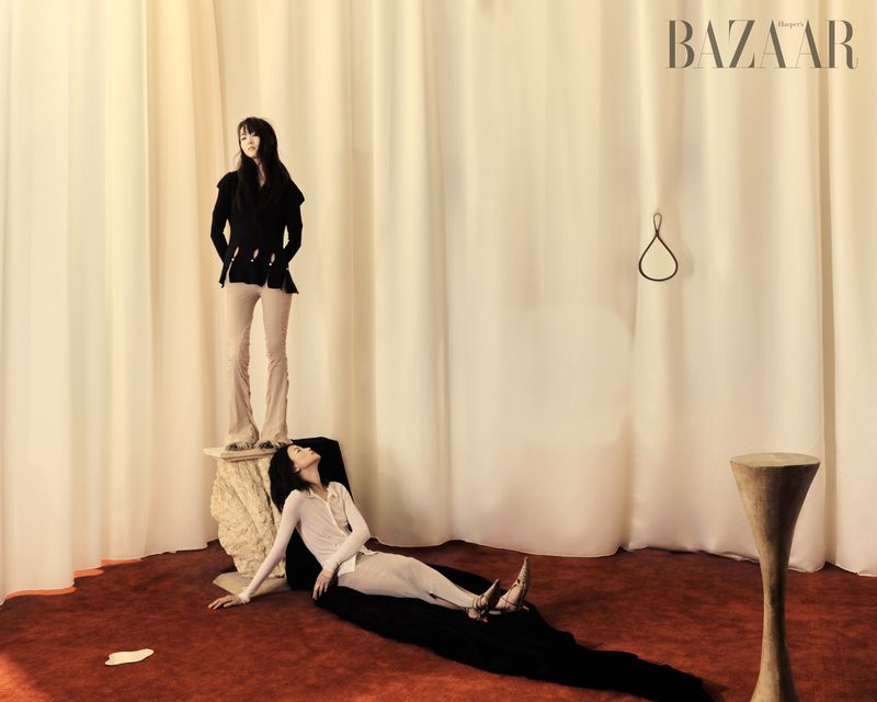 Fashion editorial shot indoors featuring two women in distinct poses, one standing on a pedestal and the other reclining on the floor.