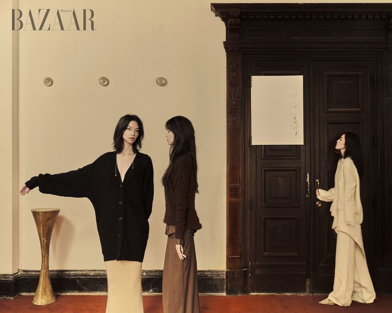 Three women in neutral clothing are posing by a dark wooden door and minimalist pedestal table in a fashion editorial shot for Bazaar.