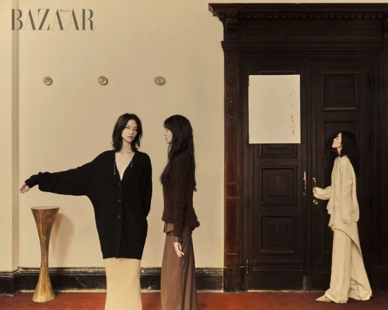 Three women in neutral clothing are posing by a dark wooden door and minimalist pedestal table in a fashion editorial shot for Bazaar.