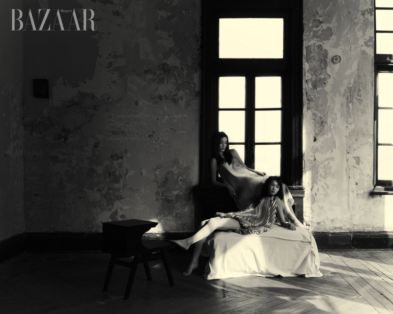 Two women in dresses pose inside a derelict interior, bathed in soft light. The setting features weathered walls, large windows, and wooden floors.
