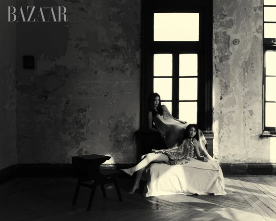 Two women in dresses pose inside a derelict interior, bathed in soft light. The setting features weathered walls, large windows, and wooden floors.