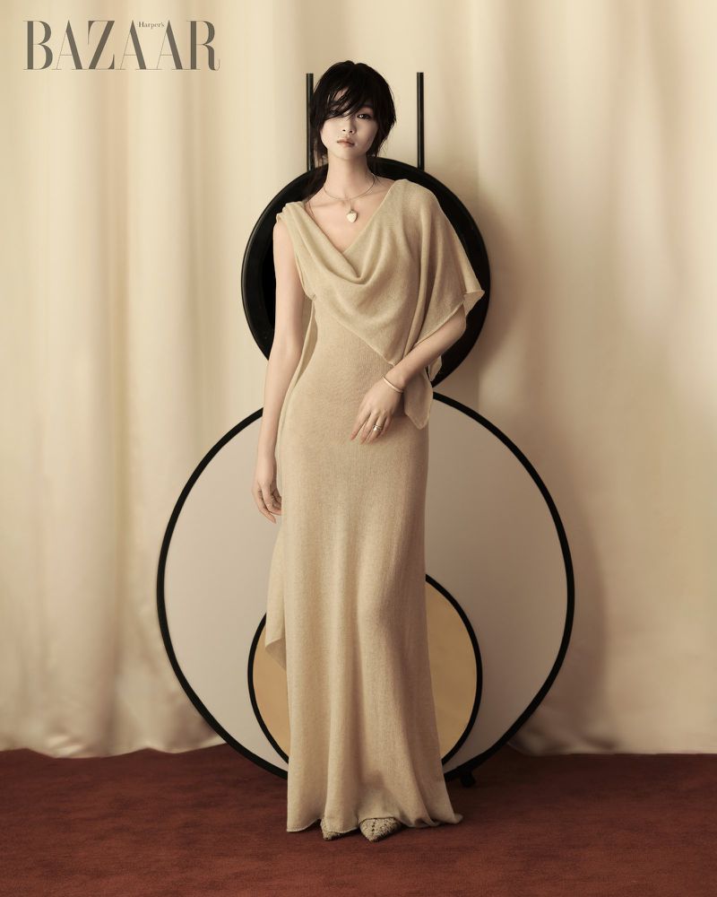 Elegant woman in a draped gown posing in front of a decorative round screen, indoors, editorial fashion photography.