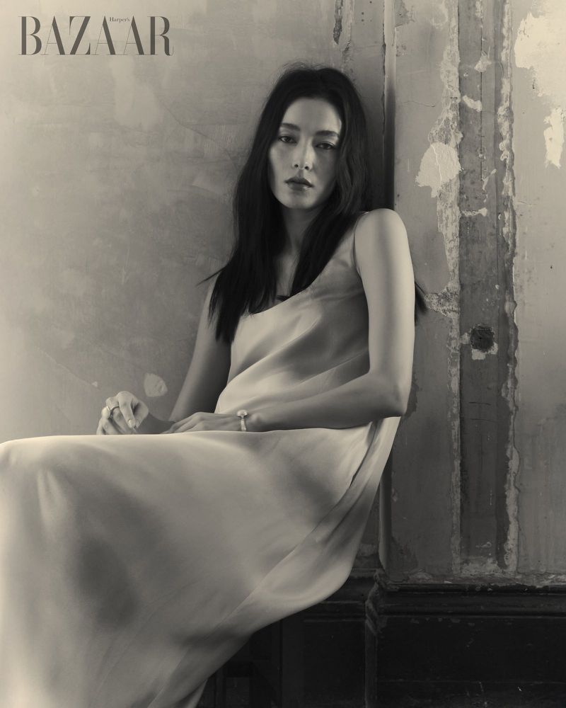 Monochrome portrait of a woman in a dress sitting against a textured wall with the Bazaar logo, creating an elegant and timeless aesthetic.