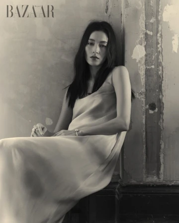 Monochrome portrait of a woman in a dress sitting against a textured wall with the Bazaar logo, creating an elegant and timeless aesthetic.