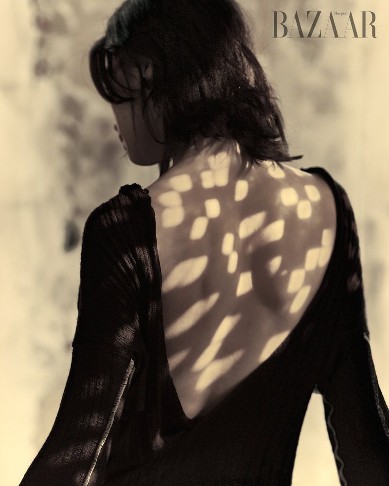Monochromatic fashion shot of a woman with dark hair wearing a black dress with a low back, posing in front of patterned lighting.
