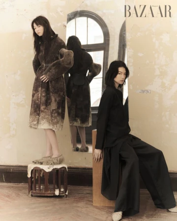 Two women pose in a sparse interior, one standing on a stool in a fur coat, the other seated in black clothing with a mirror reflecting.