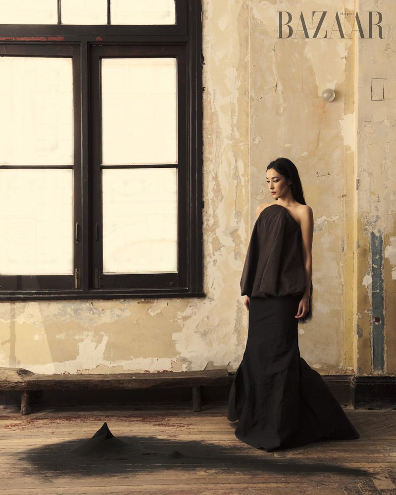 Woman in flowing brown gown stands in interior room with large window, cracked wall, and pile of dirt. Bazaar logo visible.