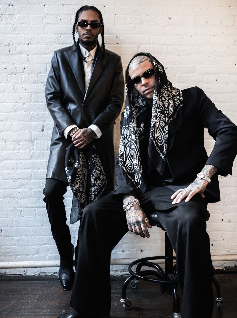 Two stylish men in black suits pose indoors against a white brick wall; one seated, both wearing sunglasses and jewelry.