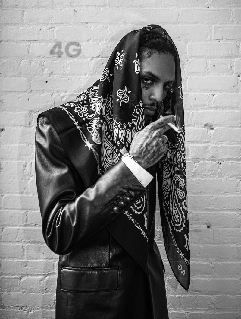 Adult male in bandana head covering and black leather jacket smokes cigarette against white brick wall in studio portrait.